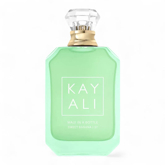 Kayali Maui in a Bottle Sweet Banana | 37 sample surrounded by banana slices, coconut shells, and tropical flowers on a sandy backdrop.