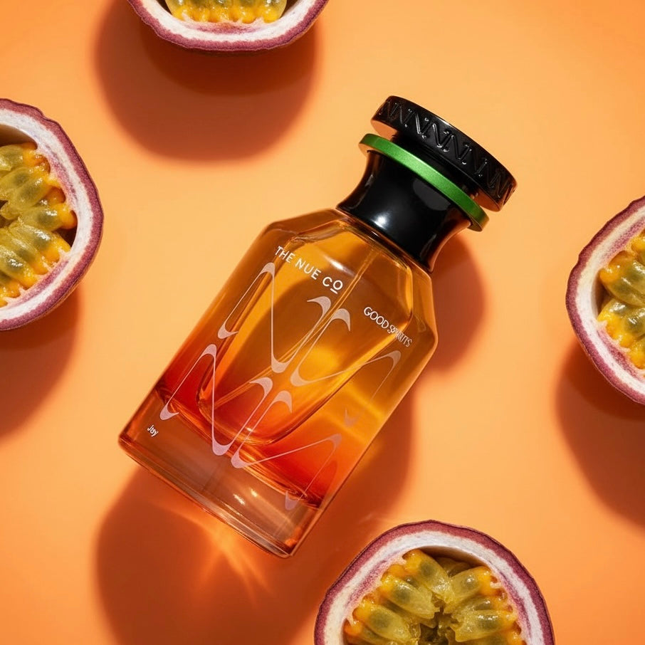 A bottle of The Nue Co Good Spirits perfume with amber-colored liquid and white graphic lettering, styled on a warm orange background with halved passion fruit slices around it.