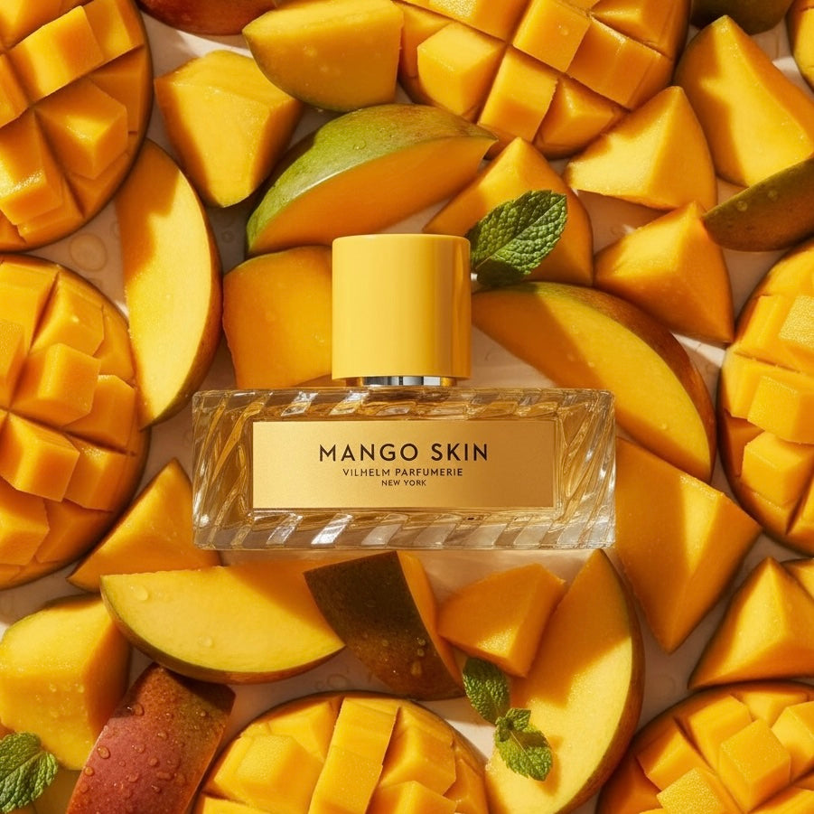 A bottle of Mango Skin perfume by Vilhelm Parfumerie lies centered on a bed of sliced and cubed mangoes with a few mint leaves. The bottle is clear glass with a yellow cap and a yellow label, surrounded by bright yellow-orange mango pieces.